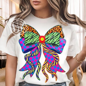 Puede incluir: Camiseta blanca con un diseño de lazo grande y colorido. El lazo incorpora varios patrones, incluyendo estampado de leopardo, rayas de cebra y un patrón de cuadros, con un detalle de botón central. La palabra "ORIGINAL" está impresa en la camiseta.