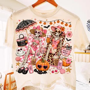 May include: Cream-colored t-shirt featuring a whimsical Halloween design. Two skeleton figures are lounging on beach chairs, surrounded by pumpkins, bats, and beach-themed accessories. The shirt includes text like "Salty Spooky & Sassy" and "Beach Witch".