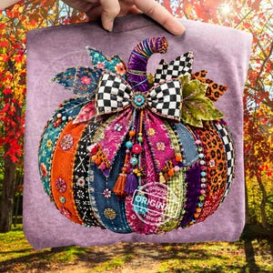 Può includere: Una felpa color viola chiaro con una grande grafica di zucca decorativa realizzata con vari ritagli di tessuto, tra cui denim, stampa leopardata e motivi floreali. La zucca è decorata con un fiocco a scacchi bianco e nero e perline.
