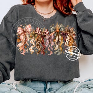May include: A charcoal gray long-sleeve sweatshirt featuring a graphic print of multiple decorative bows in various patterns including plaid, leopard print, and polka dots, accented with small pumpkins, wheat stalks, and gold stars.