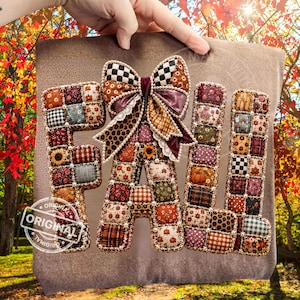 Puede incluir: Un adorno de pared que muestra la palabra FALL formada por letras acolchadas y cubiertas de tela. Cada letra está hecha con un mosaico de diversas telas con motivos otoñales, incluyendo estampados florales, de cuadros y de calabazas, decoradas con pequeñas cuentas blancas y un gran lazo de tela estampado en la parte superior de la letra A.