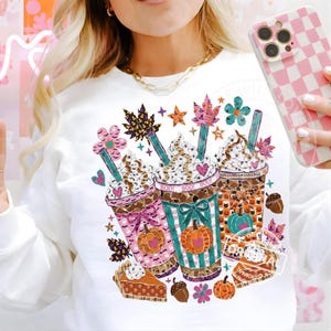 Könnte beinhalten: Ein weißes Langarm-Sweatshirt mit einem Grafikdruck von drei Eiskaffeebechern, die mit Halloween-Motiven wie Kürbissen, Geistern, Fledermäusen und Herbstblättern verziert sind. Das Design enthält bunte Strohhalme, Schlagsahne und Tortenstücke.