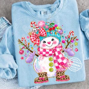 May include: A light blue crewneck sweatshirt featuring a graphic of a snowman wearing a teal hat, a pink and white checkered scarf, and leopard print boots. The snowman is decorated with a red and white checkered bow, ornaments, and lights.