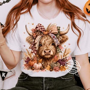 May include: A white t-shirt featuring a graphic of a Highland cow with long, shaggy brown fur. The cow is adorned with a large, multi-patterned bow between its horns and is surrounded by autumn-themed elements including pumpkins and fall leaves.