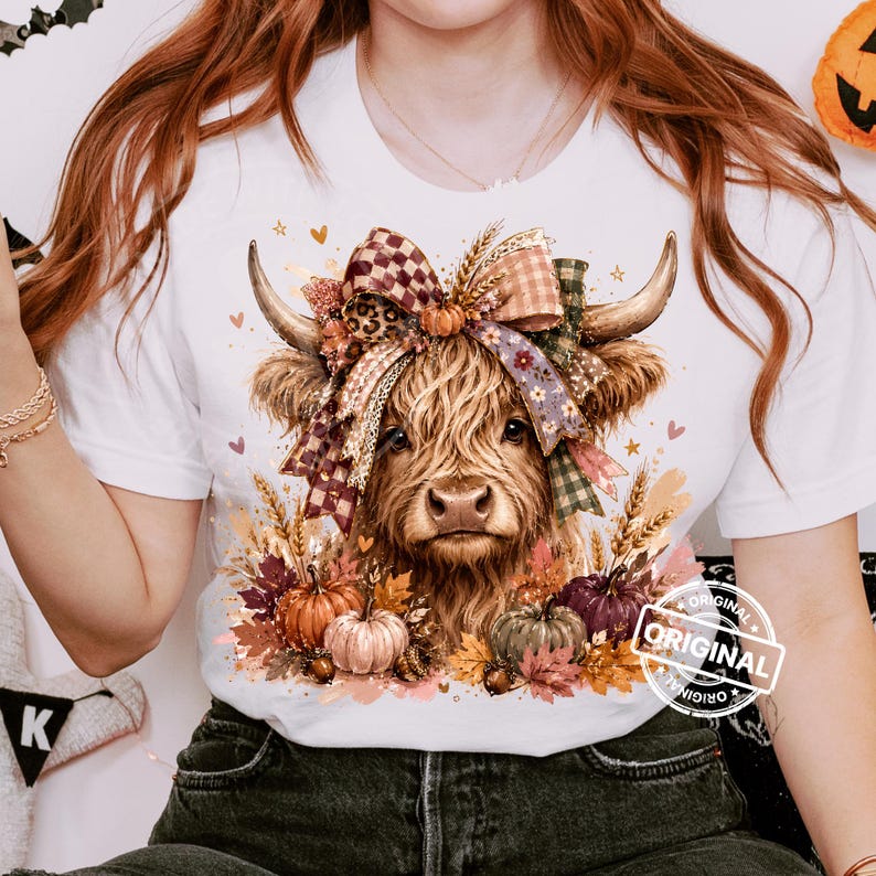 May include: A white t-shirt featuring a graphic of a Highland cow with long, shaggy brown fur. The cow is adorned with a large, multi-patterned bow between its horns and is surrounded by autumn-themed elements including pumpkins and fall leaves.