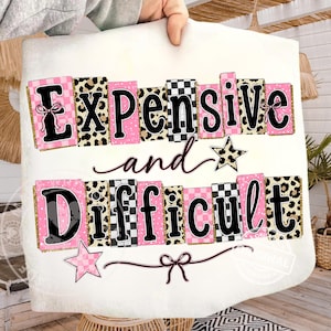 May include: White rectangular cushion featuring the phrase "Expensive and Difficult" in black, pink, and gold. The text is arranged in a stacked format with leopard print and checkered patterns. A star and bow embellish the design.
