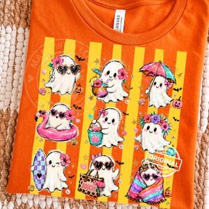 May include: An orange t-shirt featuring a graphic print of eight white ghosts in various summer activities. The ghosts are depicted with accessories like heart-shaped sunglasses, floral crowns, a pink flamingo float, a beach bag, and a colourful umbrella.