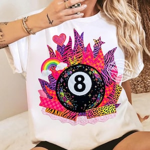 Könnte beinhalten: Weißes T-Shirt mit einem farbenfrohen Grafikdesign eines Billardkugels mit der Nummer 8, umgeben von einem lebendigen Design mit Regenbogen, Herz und Stern. Das Design enthält verschiedene Muster und Texturen, darunter Zebrastreifen und Leopardenmuster.