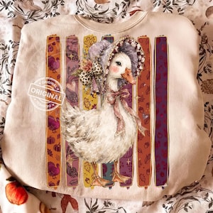 May include: A beige crewneck sweatshirt featuring a central graphic of a white goose wearing a ruffled bonnet and a pink bow. The goose is positioned in front of five vertical, colorful stripes decorated with autumn motifs like turkeys and leaves.