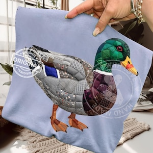May include: A decorative pillow featuring a detailed patchwork design of a mallard duck. The duck has a green head, a yellow beak, and a body composed of various fabric patterns. The pillow is held against a light blue background.
