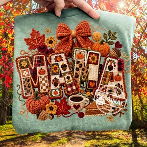 Puede incluir: Una sudadera verde claro con un gran diseño bordado de múltiples texturas que forma la palabra MAMA. Las letras están decoradas con motivos otoñales como calabazas, girasoles, pavos y cuadros, acentuadas por un lazo y una taza de ganchillo.
