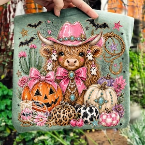May include: A green tote bag featuring a whimsical illustration of a Highland cow wearing a pink cowboy hat, surrounded by pumpkins, bats, and Halloween-themed decorations. The design includes the text "ORIGINAL".