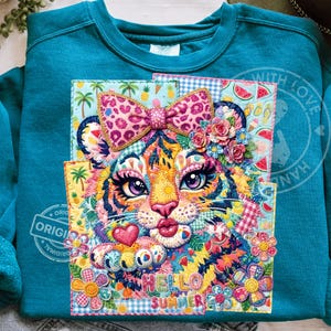 Peut inclure: Sweat-shirt teal avec un motif appliqué de visage de tigre coloré. Le tigre a un nœud rose, des fleurs et les mots "HELLO SUMMER". Le motif comprend des palmiers, des ananas et des tranches de pastèque.