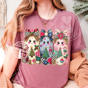 May include: A mauve t-shirt featuring a graphic of three stylized ghosts decorated with Christmas elements. Each ghost wears a large bow and displays patterns including plaid, leopard print, and polka dots, alongside images of trees and candy canes.