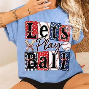 May include: Light blue t-shirt with the words "Let's Play Ball" in a baseball-themed design. The design incorporates baseballs, bats, and patterns in red, white, and blue.