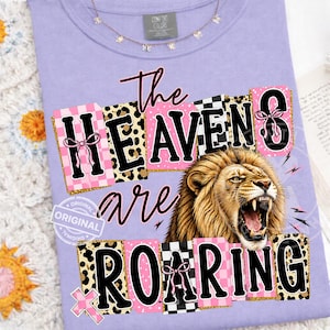 Op de afbeelding: Een lila Comfort Colors T-shirt met de tekst "The Heavens are Roaring" en een brullende leeuw. De tekst bevat verschillende patronen, waaronder luipaardprint en roze ruitjes.
