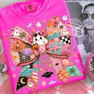 May include: A hot pink t-shirt featuring a large, colorful bow design. The bow is decorated with various patches including a ghost wearing sunglasses, pumpkins, seashells, and stars. The design has a patchwork aesthetic with gold trim and a playful, whimsical theme.