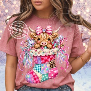 May include: A mauve t-shirt featuring a graphic of a fluffy Highland cow with a pink and white striped bow, peeking out of a colorful patchwork Christmas stocking decorated with disco balls, candy canes, and festive lights. The text ORIGINAL is visible.
