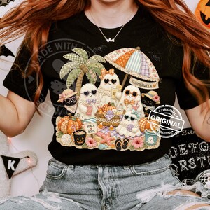 May include: Black t-shirt featuring a whimsical "Summerween" design. The graphic includes ghost characters at a beach, with pumpkins, a palm tree, and an umbrella. The text "Beach Boos" is also visible.