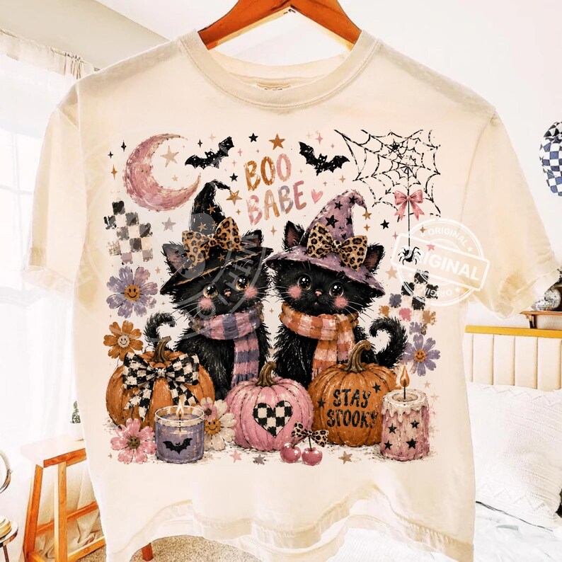 May include: Cream-colored t-shirt featuring a Halloween-themed graphic. Two black cats wearing witch hats and scarves are surrounded by pumpkins, candles, and floral accents. The text "BOO BABE" and "STAY SPOOKY" are also visible.