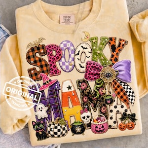 May include: A pale yellow crewneck sweatshirt featuring the text SPOOKY MAMA in large, patterned letters. The design includes Halloween motifs like a witch hat, ghost, black cat, pumpkin, skull, cauldron, spider web, and a purple bow.