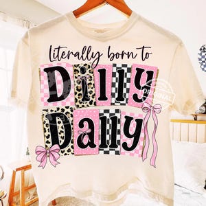 Könnte beinhalten: Cremefarbenes T-Shirt mit dem Schriftzug "Literally born to Dilly Dally" in schwarzer Schrift. Der Text ist über Blöcken mit rosa Karomustern, Leopardenmuster und Polka-Dots angeordnet. Rosa Schleifen akzentuieren das Design.