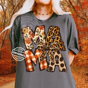 Puede incluir: Una camiseta gris carb&oacute;n con la palabra MAMA en letras grandes. Cada letra tiene un patr&oacute;n diferente: floral, estampado de leopardo, cuadros naranjas y lunares marrones. Un lazo con estampado de leopardo est&aacute; centrado sobre la letra M.