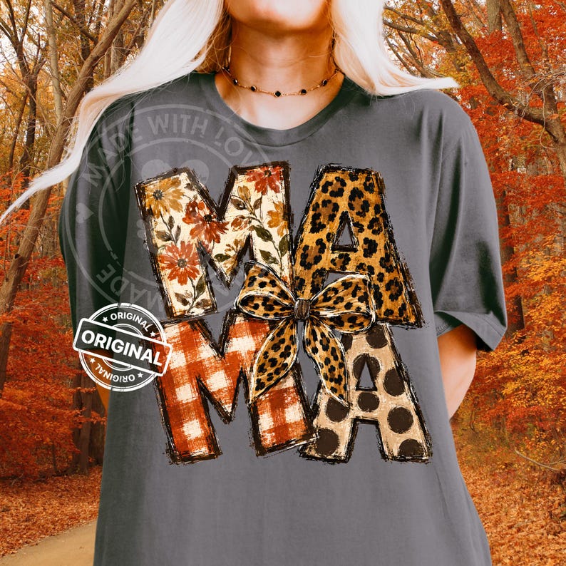 Puede incluir: Una camiseta gris carb&oacute;n con la palabra MAMA en letras grandes. Cada letra tiene un patr&oacute;n diferente: floral, estampado de leopardo, cuadros naranjas y lunares marrones. Un lazo con estampado de leopardo est&aacute; centrado sobre la letra M.