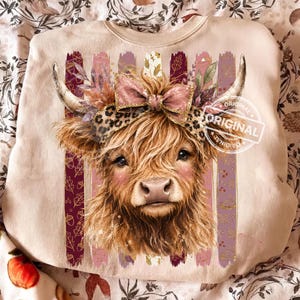 May include: A beige crewneck sweatshirt featuring a graphic of a fluffy Highland cow wearing a leopard print headband with a large bow. The cow is centered against vertical stripes decorated with autumn leaves and acorns in shades of pink and brown.