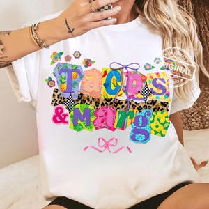 May include: White t-shirt with the text "Tacos & Margs" in colorful, patchwork-style letters. The design includes floral and animal print accents, with a pink bow at the bottom. The shirt is a casual, fun style.