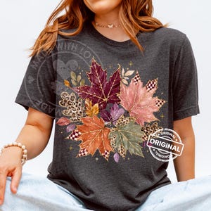 Può includere: Una t-shirt grigio antracite scuro con una grafica centrale di varie foglie d'acero autunnali nei toni del bordeaux, arancione, rosa e verde. Le foglie sono decorate con accenti glitter dorati, motivi leopardati e piccoli quadrati a scacchi.