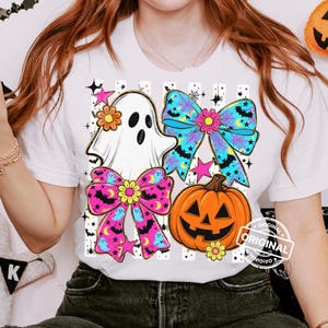 以下が含まれることがあります： ハロウィーンをテーマにしたデザインの白いTシャツ。デザインには、お化け、コウモリの付いた2つのリボン、カボチャ、星が含まれています。リボンはピンクと青で、黄色の花のアクセントが付いています。「ORIGINAL」の文字がプリントされています。