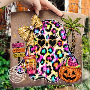 May include: A brown t-shirt featuring a graphic of a ghost with a colourful leopard print pattern, wearing heart-shaped sunglasses and a gold glitter bow. The design includes a palm tree, a pumpkin bucket with a watermelon slice, and a signpost reading BEACH, BOO, and BOO BEACH, alongside a cup labelled too cute TO SPOOK.