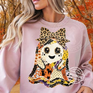 May include: A light pink sweatshirt featuring a central graphic of a ghost wearing a leopard print bow. The ghost is composed of various patterned patches including pumpkins, skulls, black cats, and spider webs in orange, black, and white.