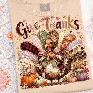 May include: A beige t-shirt featuring a graphic of a turkey with a fan of colorful patterned feathers, including plaid, leopard print, and floral designs. The text "Give Thanks" is printed above the turkey, surrounded by pumpkins and wheat.