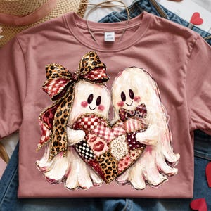 May include: A dusty rose t-shirt featuring a graphic of two white ghosts holding a large heart. The left ghost wears a large leopard print and red plaid bow, while the right ghost wears a red plaid bowtie. The heart has a patchwork pattern of red, white, and black designs including lips, hearts, and leopard print.