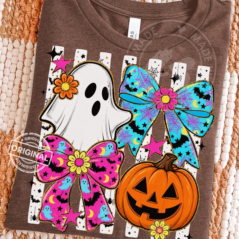 以下が含まれることがあります： ハロウィーンをテーマにしたデザインの茶色のTシャツ。白い幽霊、コウモリと月のある2つのカラフルなリボン、ジャック・オー・ランタン、星が含まれています。リボンはピンクと青のアクセントがあり、幽霊には花が付いています。
