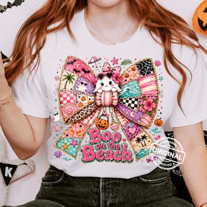 May include: White t-shirt featuring a colorful bow design with a ghost character wearing sunglasses. The bow is made of patchwork patterns with beach and Halloween elements. Text reads "Boo on the Beach".