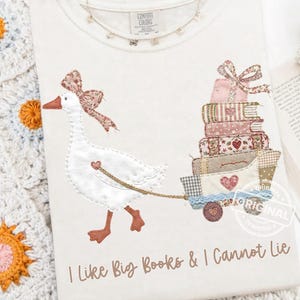 Puede incluir: Camiseta color crema con un dise&ntilde;o caprichoso de un ganso tirando de un carro lleno de libros. El ganso tiene un lazo rosa y tira de un carro con una pila de libros. El texto "I Like Big Books & I Cannot Lie" est&aacute; impreso debajo.