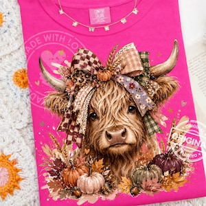 May include: A bright pink t-shirt featuring a graphic of a Highland cow with a large, multi-patterned bow between its horns. The design includes pumpkins, autumn leaves, and wheat stalks at the base. The inner neck label reads Comfort Colors.