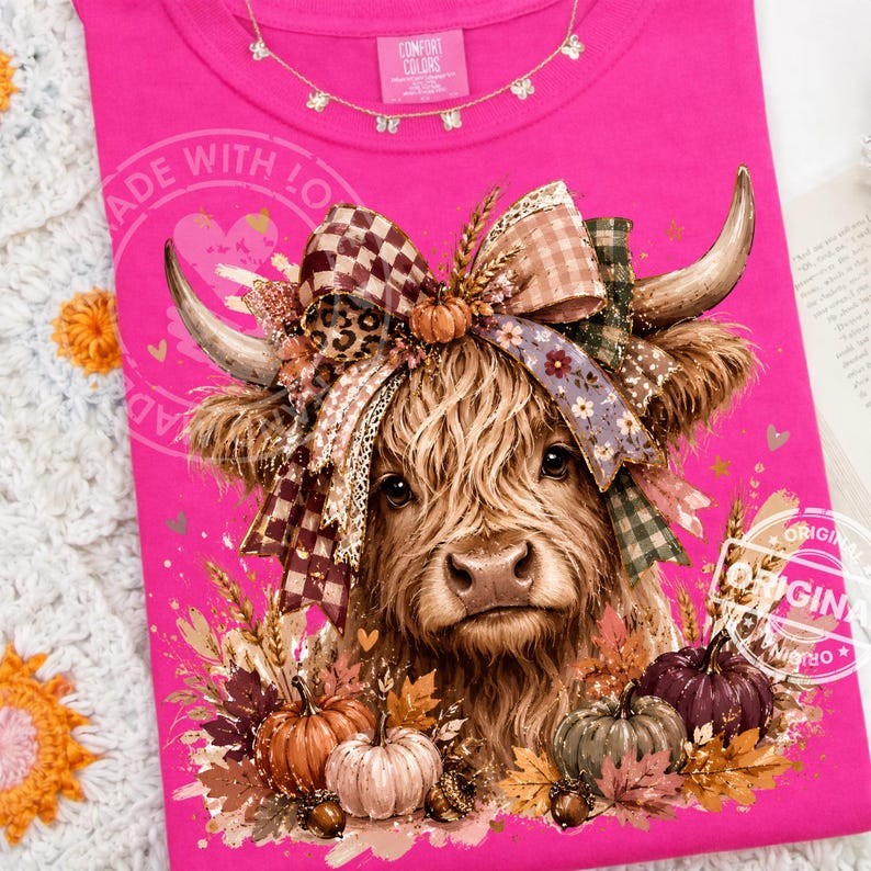 May include: A bright pink t-shirt featuring a graphic of a Highland cow with a large, multi-patterned bow between its horns. The design includes pumpkins, autumn leaves, and wheat stalks at the base. The inner neck label reads Comfort Colors.