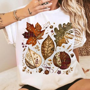 May include: A white t-shirt featuring a graphic print of various autumn leaves, including maple and oak shapes, with plaid and textured patterns. The design includes small mushrooms, acorns, berries, and a wax seal stamp with the text ORIGINAL.