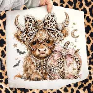 May include: A white fabric transfer featuring a fluffy brown Highland cow wearing heart-shaped leopard print glasses, a large leopard print bow, and a pearl necklace. Beside the cow is a pumpkin with a pink bow, spiderwebs, bats, and stars.