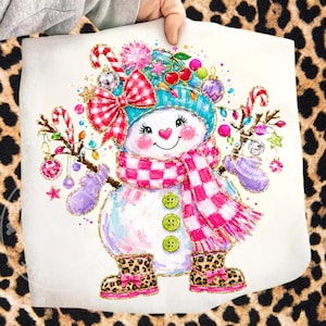 May include: A white snowman graphic print featuring a teal hat, a pink and white checkered scarf, and three green buttons. The snowman wears leopard print boots with pink bows and holds candy canes and ornaments on brown branch arms.