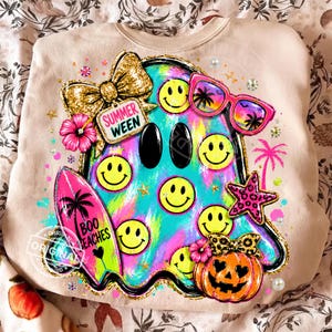 Op de afbeelding: Een beige sweatshirt met een kleurrijke afbeelding van een geest bedekt met gele smileys. Het ontwerp bevat een gouden glitterstrik, een roze zonnebril, een surfplank met de tekst "BOO SEACHES", een pompoen en de tekst "SUMMER WEEN".