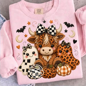 May include: A light pink long-sleeve sweatshirt featuring a central graphic of a brown highland cow with a black and white checkered bow, flanked by a white ghost with a checkered pattern and an orange leopard print ghost, plus two pumpkins.