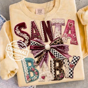 Puede incluir: Una sudadera de cuello redondo en color amarillo pálido con el texto SANTA BABY en letras grandes y decorativas. Las letras están adornadas con bordados florales, encaje, estampado de leopardo y cuadros, con un gran lazo texturizado central.