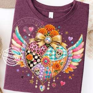 Op de afbeelding: Een bordeauxrood t-shirt met een centrale afbeelding van een hart met vleugels, versierd met luipaardprint, ruitpatronen, suikerschedels, oranje goudsbloemen en een gouden strik. Het ontwerp bevat kleurrijke strass-steentjes en hangende bedels.