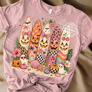 May include: Pink t-shirt featuring a Halloween-themed design. The design includes five surfboards with jack-o'-lantern faces, bows, and various patterns. Pumpkins, ghosts, and seashells are also part of the design.