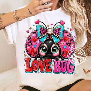 Pode incluir: Uma t-shirt branca com o gráfico de uma joaninha preta de olhos grandes, usando um laço azul brilhante e rosa com estampa de leopardo. O design inclui corações vermelhos, raios azuis, estrelas douradas e o texto LOVE BUG em letras.